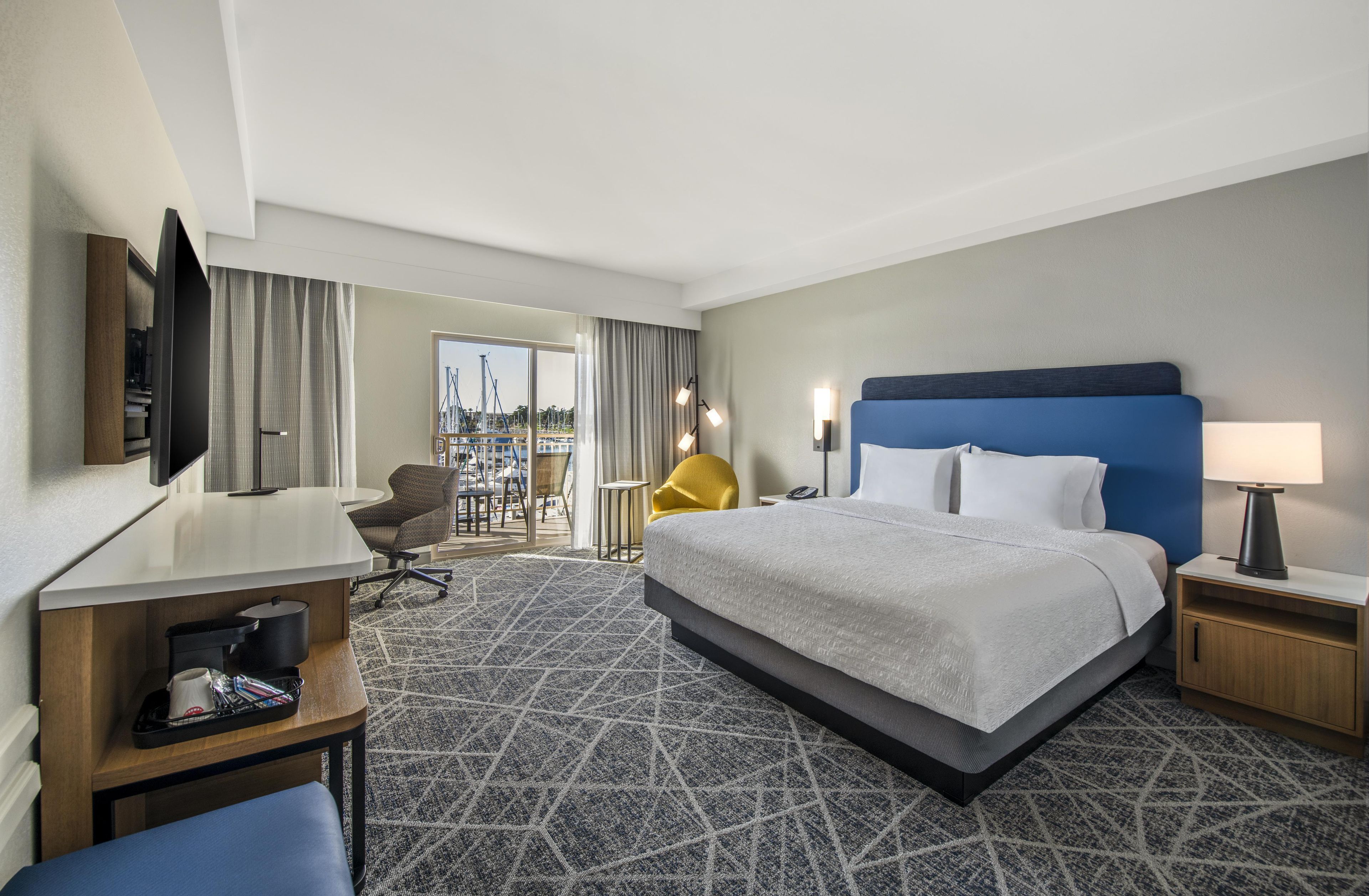 A hotel room with a bed, a desk, a chair, a television, and a view of the city.