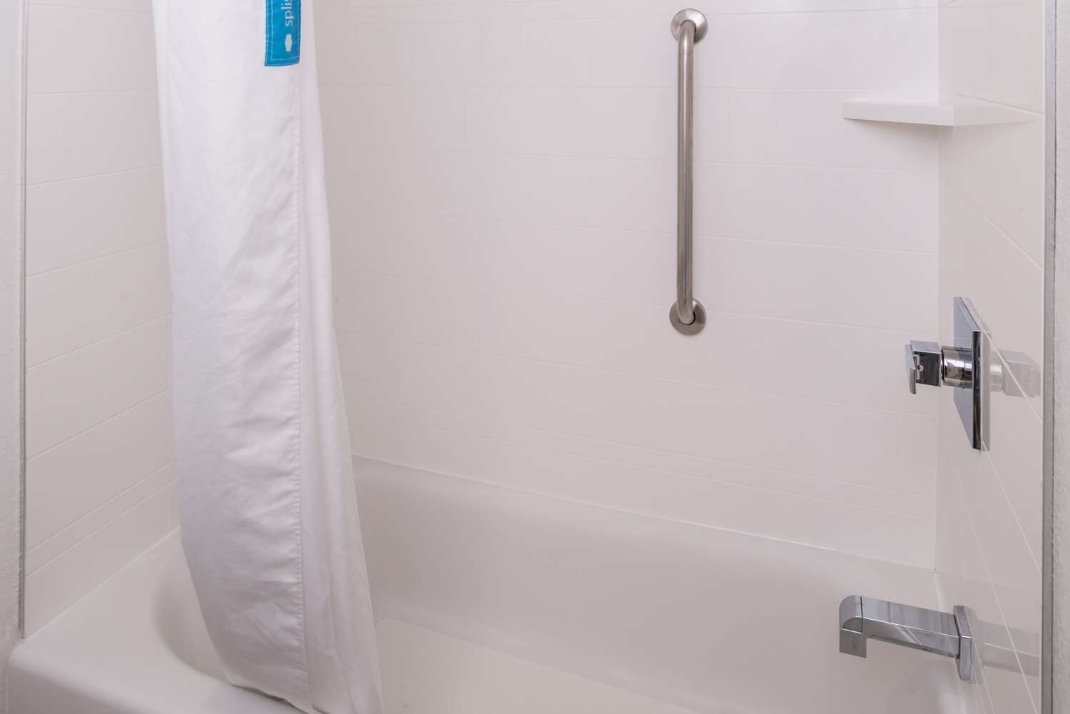 Un espace douche équipé d’un rideau de douche blanc, d’une barre d’appui et d’un pommeau de douche à main.