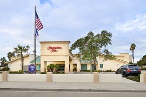 A Hampton Inn hotel with a beige facade, American flags, and palm trees. - Hampton Inn Channel Islands Harbor/Oxnard (Oxnard)