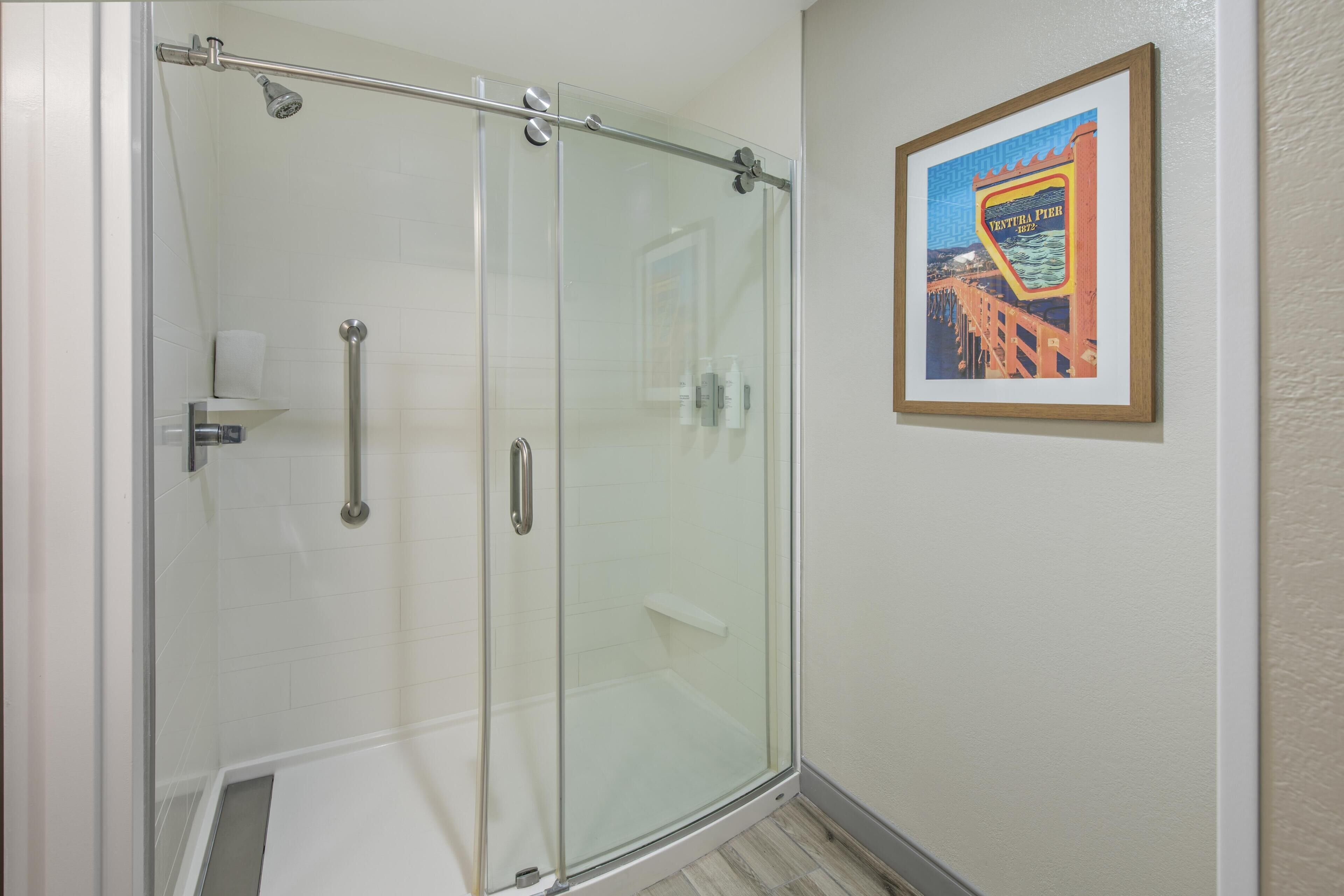 A glass-enclosed shower with a handrail, a framed picture on the wall, and a white tiled floor.