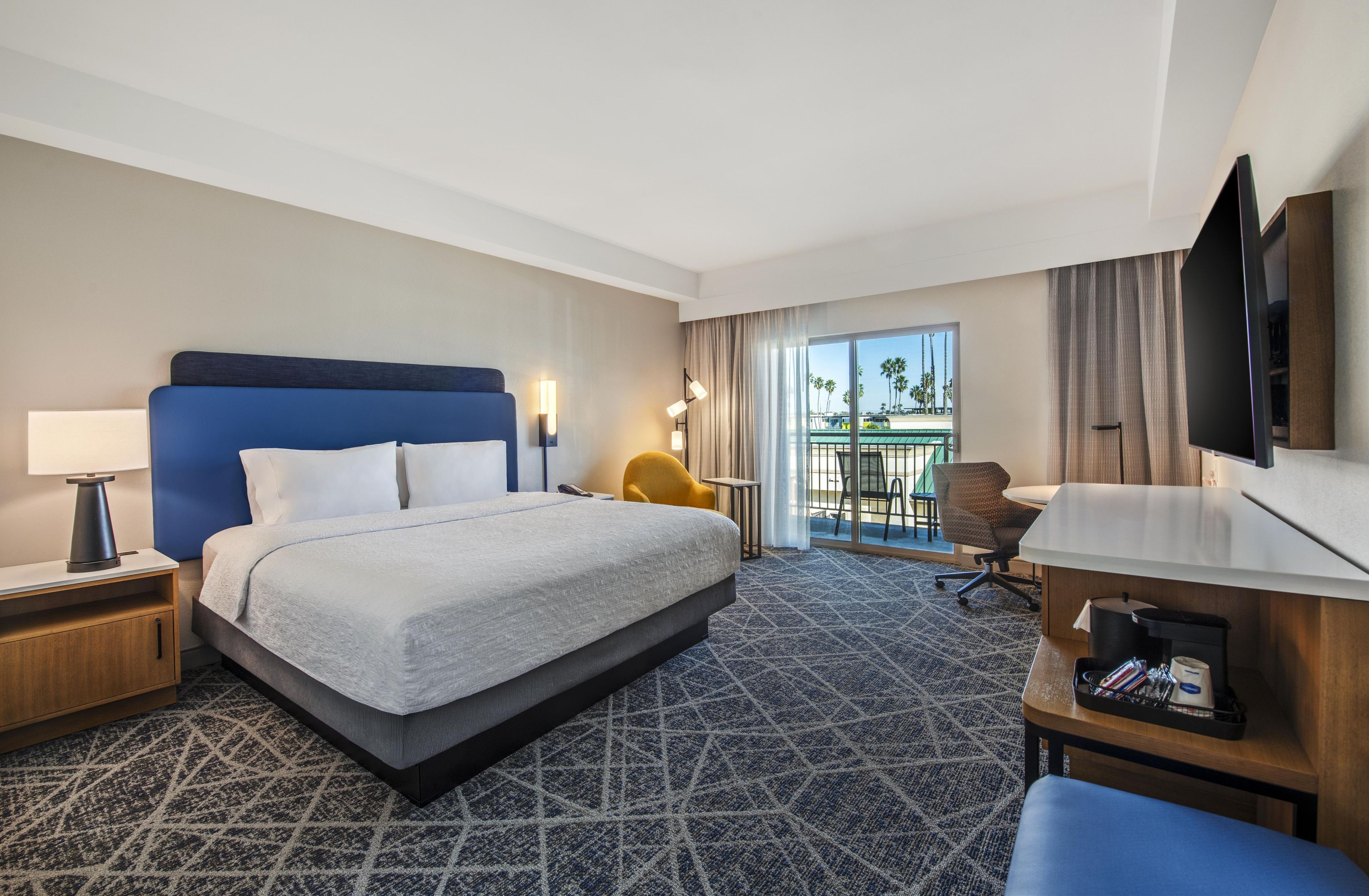 Habitación de hotel con una cama grande, un escritorio, una silla, un televisor y un balcón con vistas a palmeras.