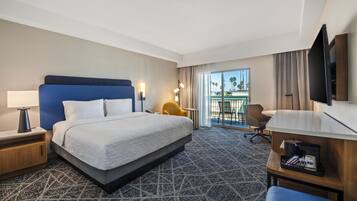 Habitación de hotel con una cama grande, un escritorio, una silla, un televisor y un balcón con vistas a palmeras.