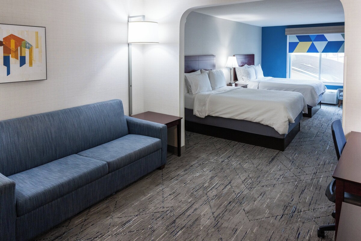 Quarto de hotel com um sofá azul, duas camas, uma escrivaninha e uma luminária.
