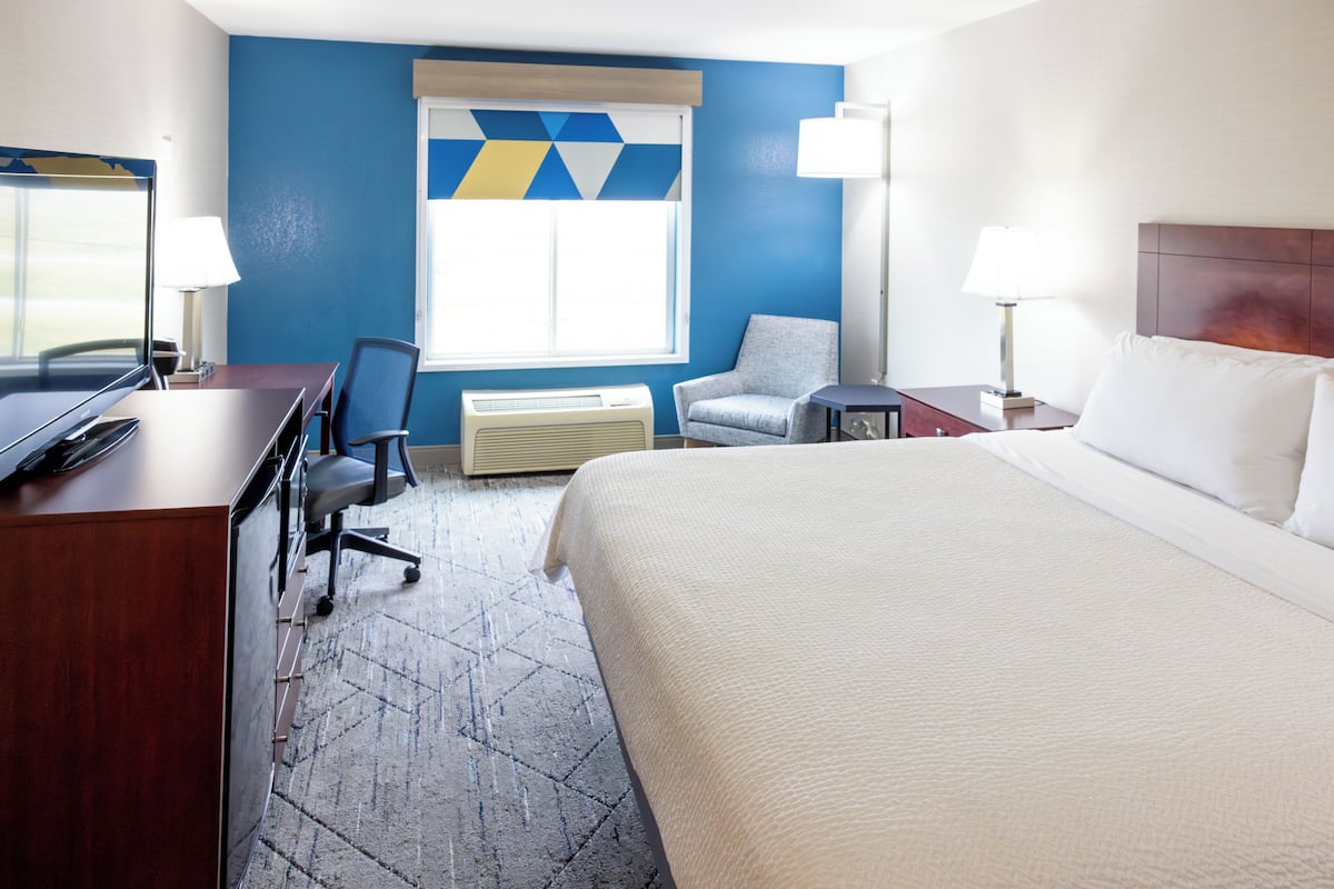 Ein Hotelzimmer mit einem großen Bett, einem Schreibtisch mit Computer, einem Sessel, einem Fenster mit Jalousien und einer Wand mit geometrischem Muster.