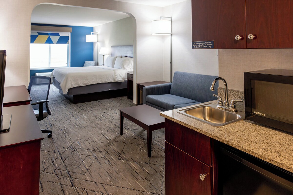 A compact hotel room with a bed, a sofa, a small table, a kitchenette with a sink, and a television.