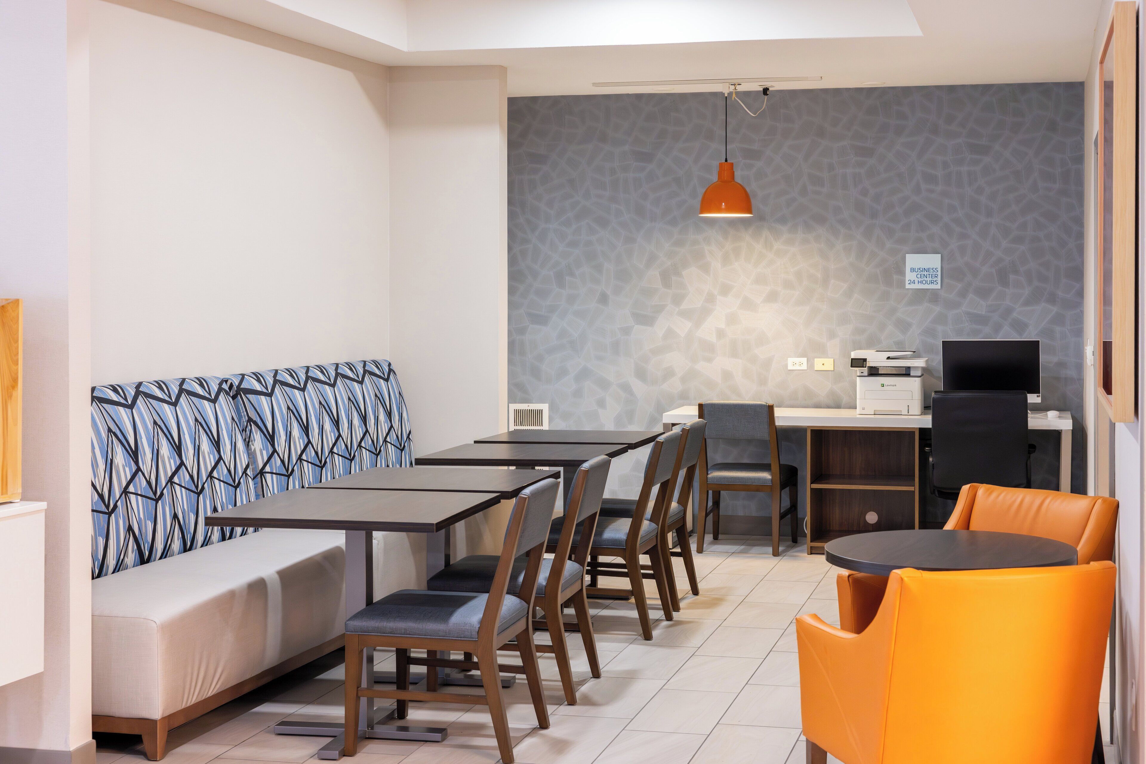 A modern dining area with a long table, multiple chairs, a sofa, and a small kitchen area with a printer and monitor.