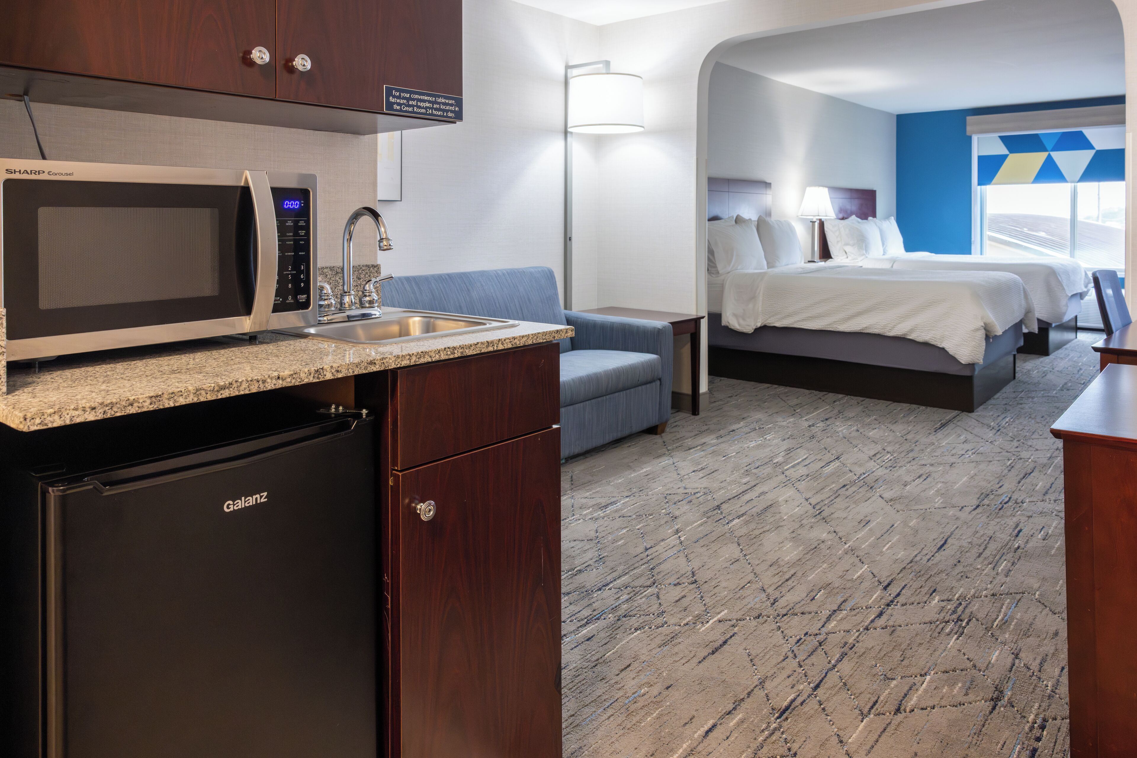 A compact hotel room with a kitchenette, a bed, a sofa, and a view of the outside.