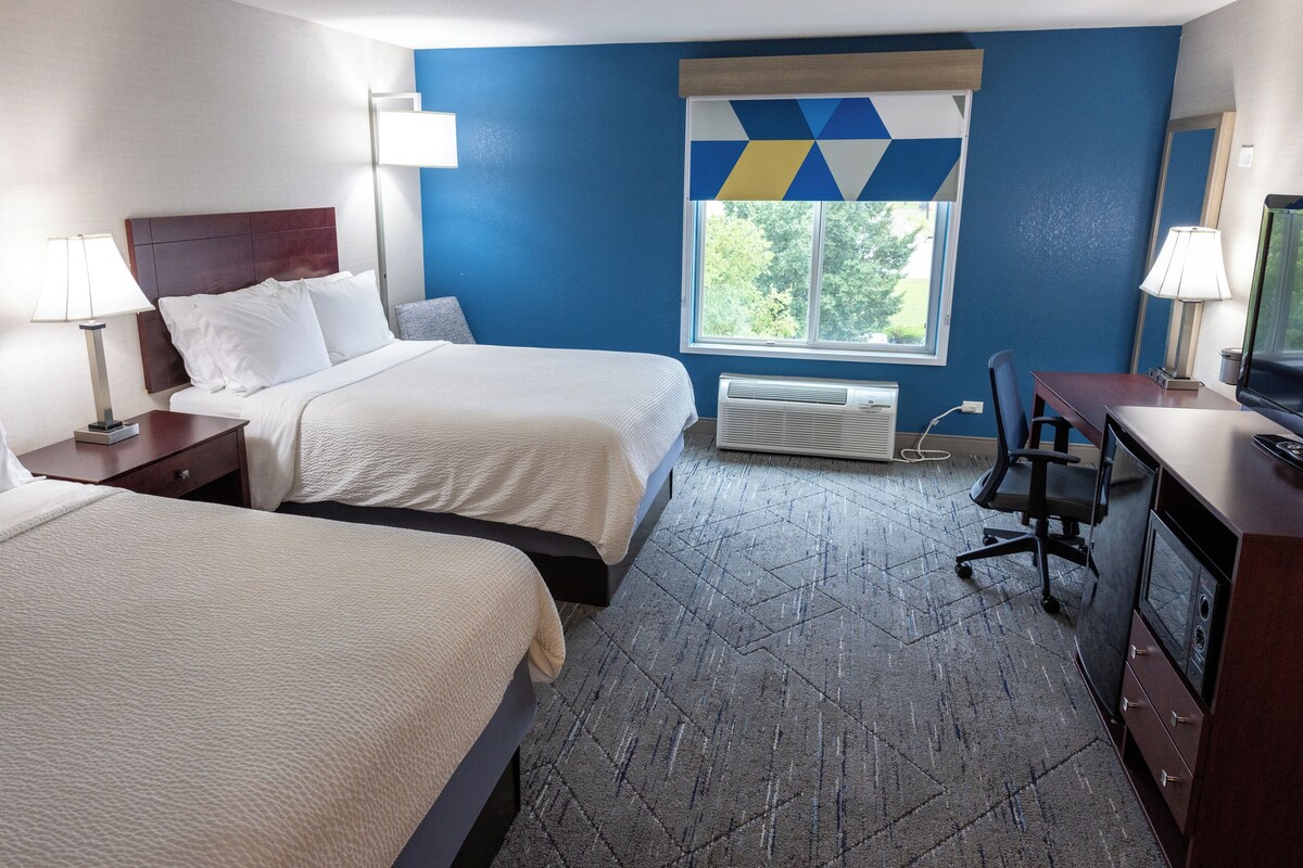 A hotel room with two beds, a desk, a chair, a television, and a window with a view of trees.