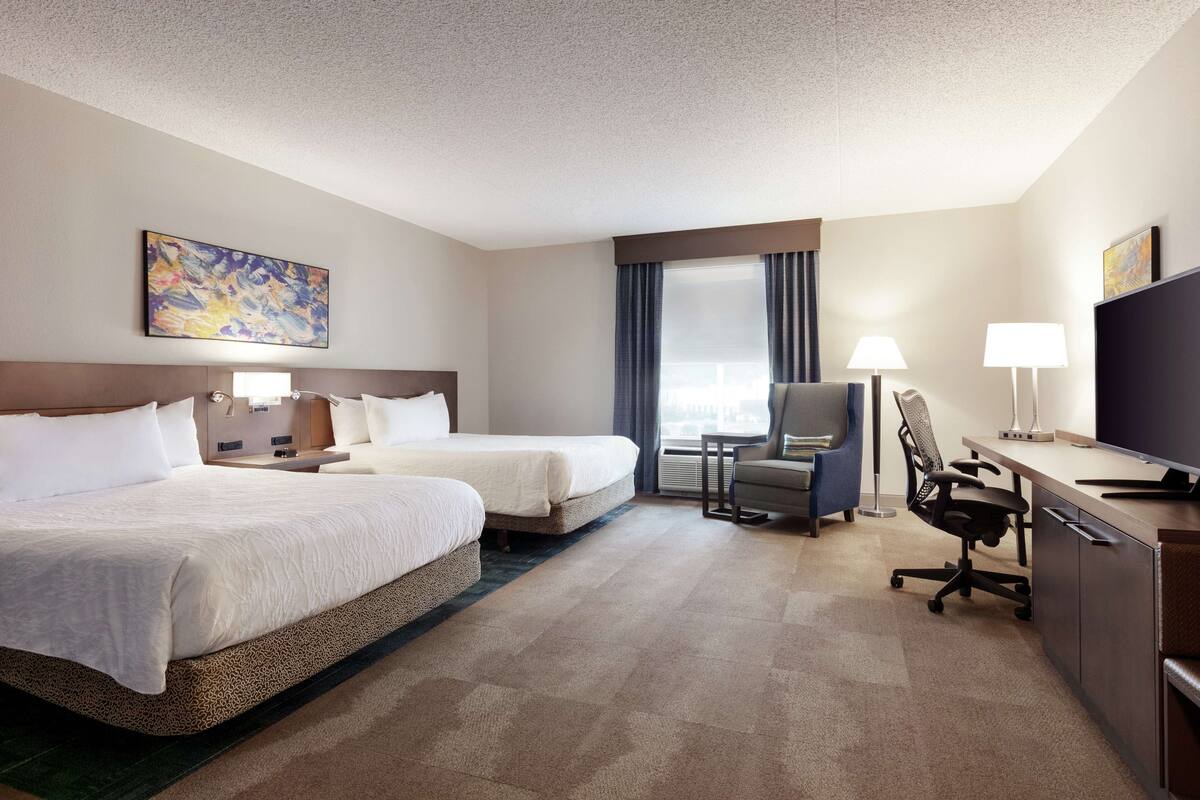 Une chambre d’hôtel avec deux lits, un bureau avec un ordinateur, une chaise, une télévision et un tableau au mur.