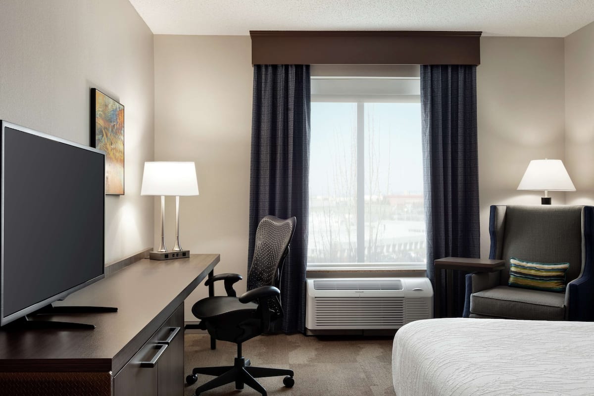 Une chambre d’hôtel comprenant un lit, un bureau avec un téléviseur à écran plat, une chaise, une lampe et une fenêtre avec des rideaux.