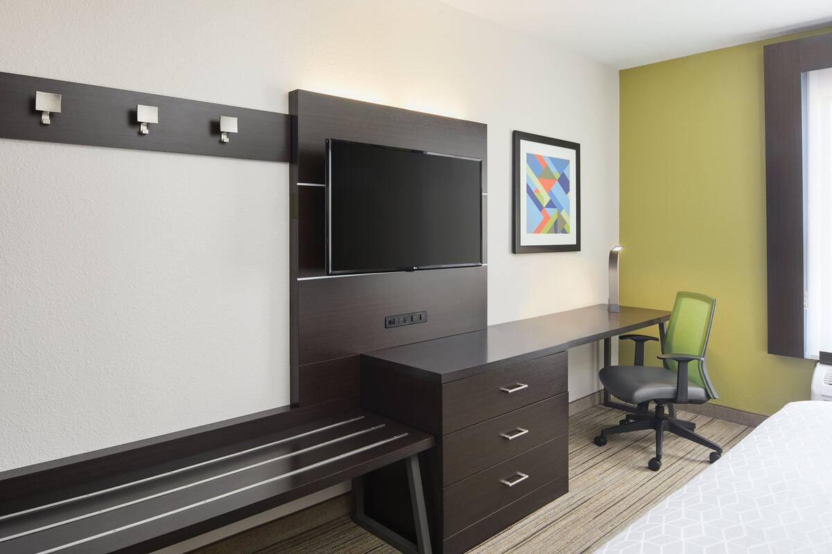 A modern hotel room with a dark wooden desk, a flat-screen TV, a green office chair, and a bed with a patterned bedspread.