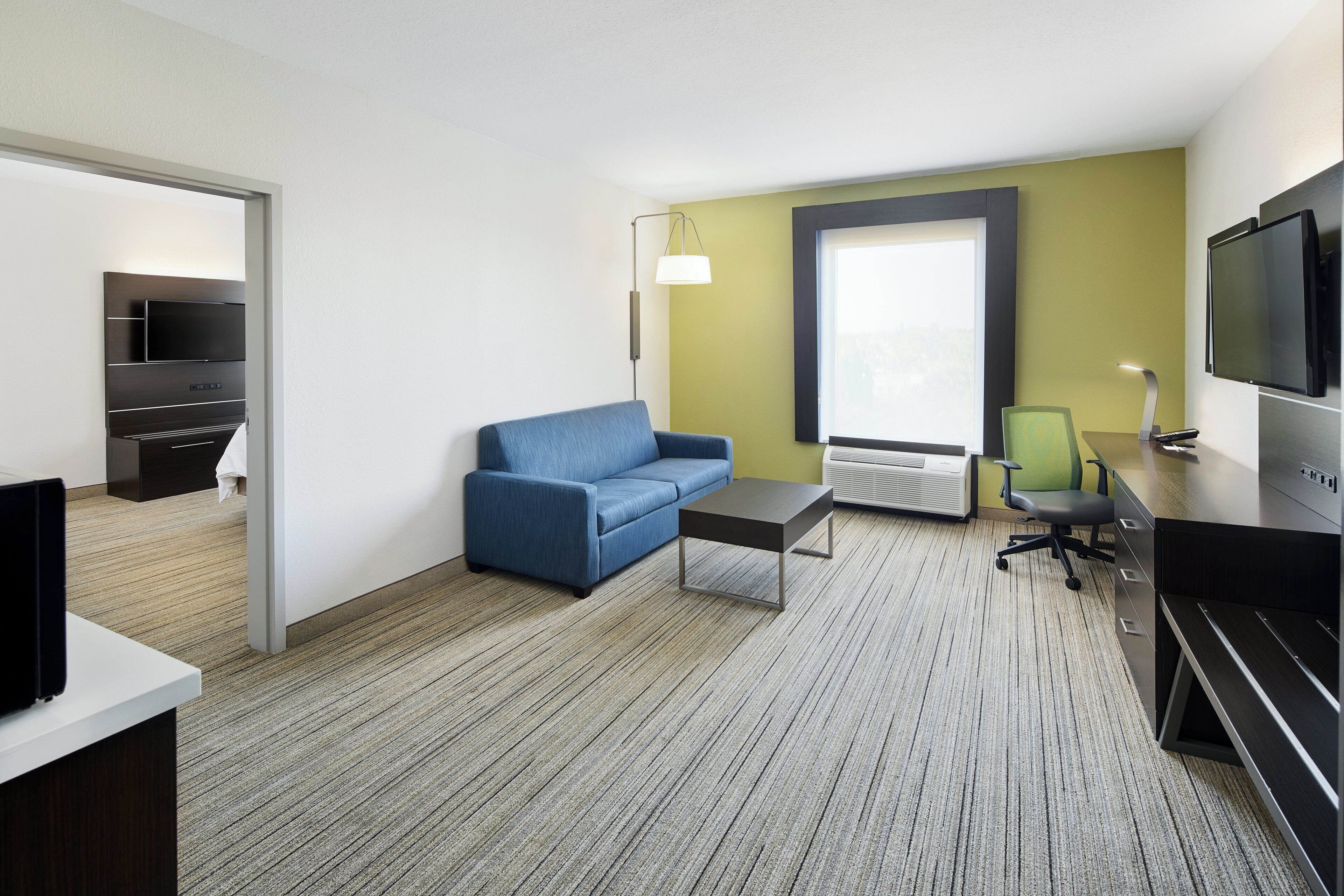 Ein Hotelzimmer mit einer Couch, einem kleinen Tisch, einem Fernseher, einem Schreibtisch mit Computer und einem Fenster mit Blick.