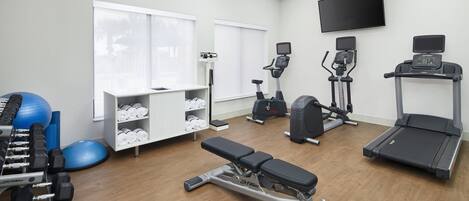 Ein Fitnessraum mit Trainingsgeräten, einer Bank, einem Laufband und einem an der Wand befestigten Fernseher.