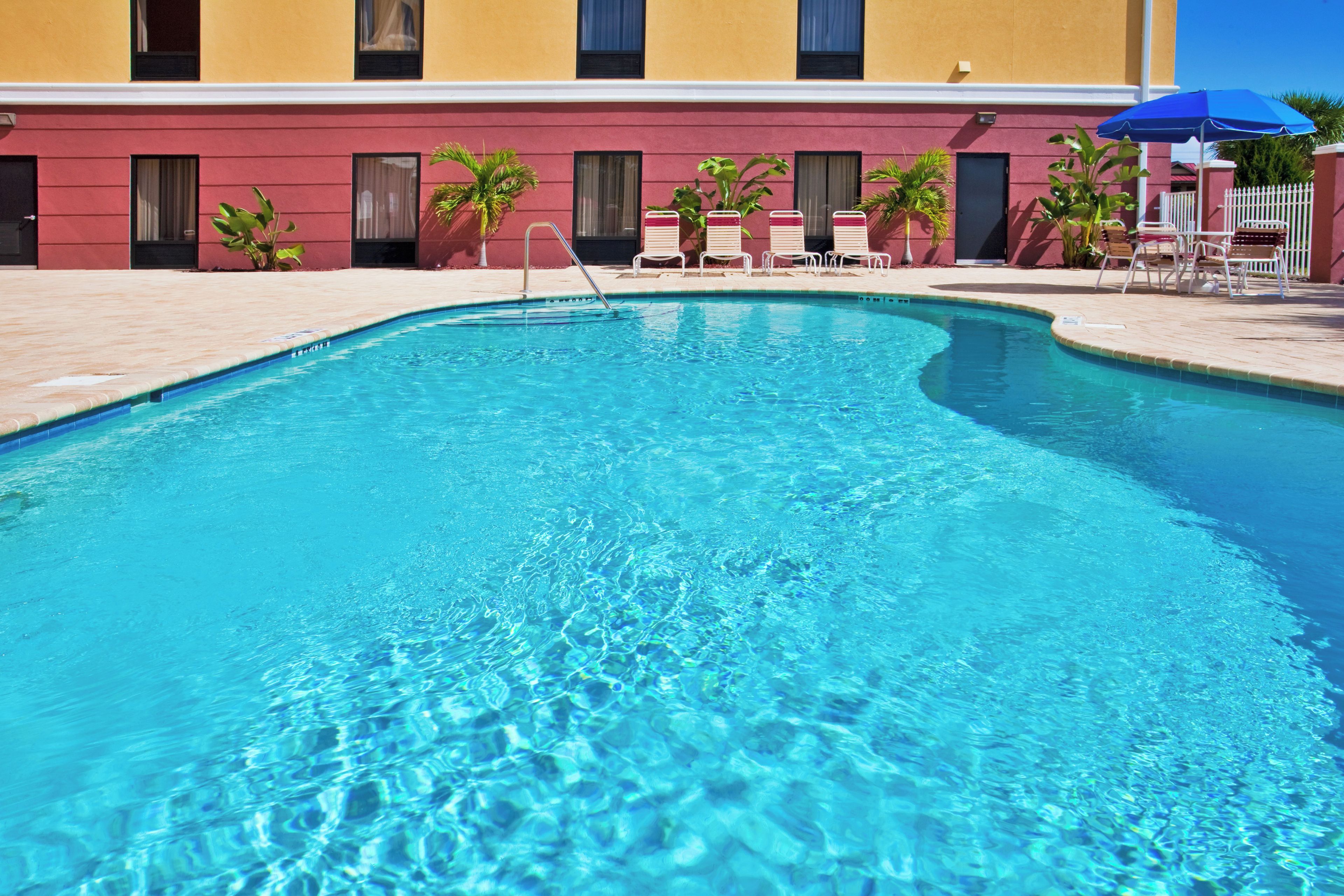 A clear blue pool with a pool ladder, surrounded by lounge chairs and umbrellas, with a building in the background.