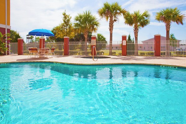 A clear swimming pool with a fenced area, palm trees, and a blue umbrella.