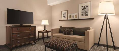 A hotel room with a television, a sofa, an ottoman, a lamp, and framed artwork on the wall.