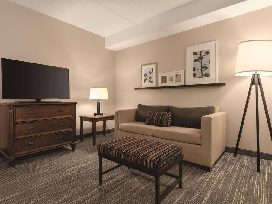 Quarto de hotel com televisão, sofá, banquinho, luminária e obras de arte emolduradas na parede.