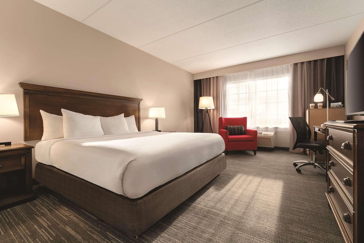 A hotel room with a large bed, two bedside lamps, a red armchair, a desk with a lamp, and a dresser.