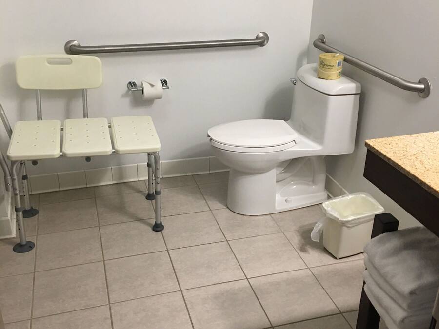A bathroom with a wheelchair-accessible toilet, a handrail, a sink, and a small trash bin.