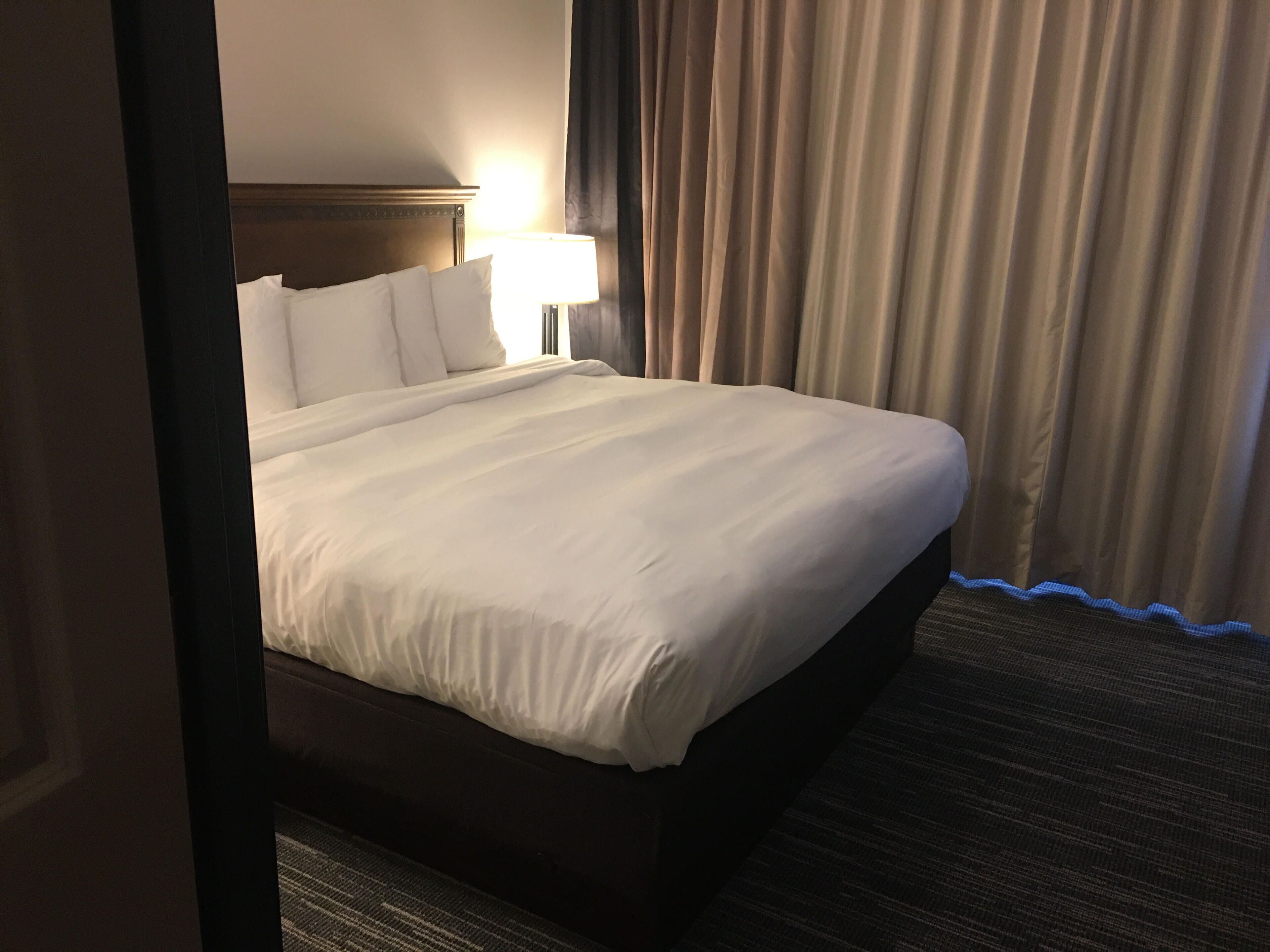 Habitación de hotel con una cama grande, una mesita de noche con lámpara y vistas al exterior a través de cortinas.
