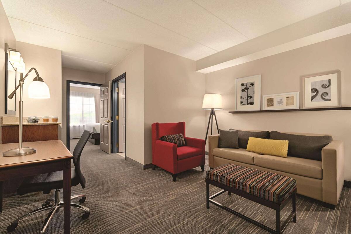 A hotel room with a desk, office chair, red armchair, sofa, and a small ottoman.