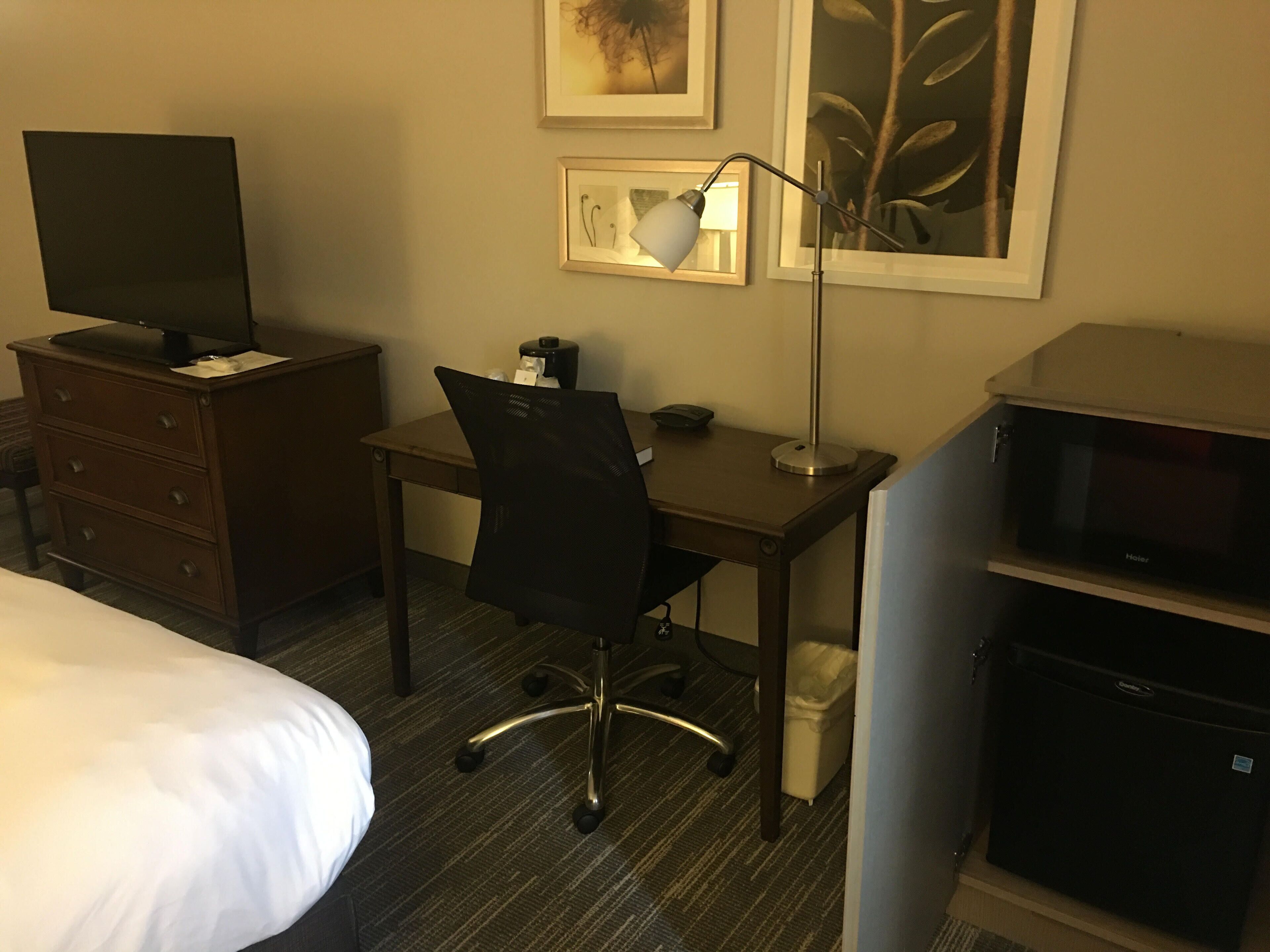 A hotel room with a bed, a desk with a computer, a chair, a lamp, a television, and a minibar.