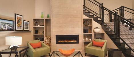 A modern living room with a fireplace, green armchairs, a glass coffee table, and a staircase.