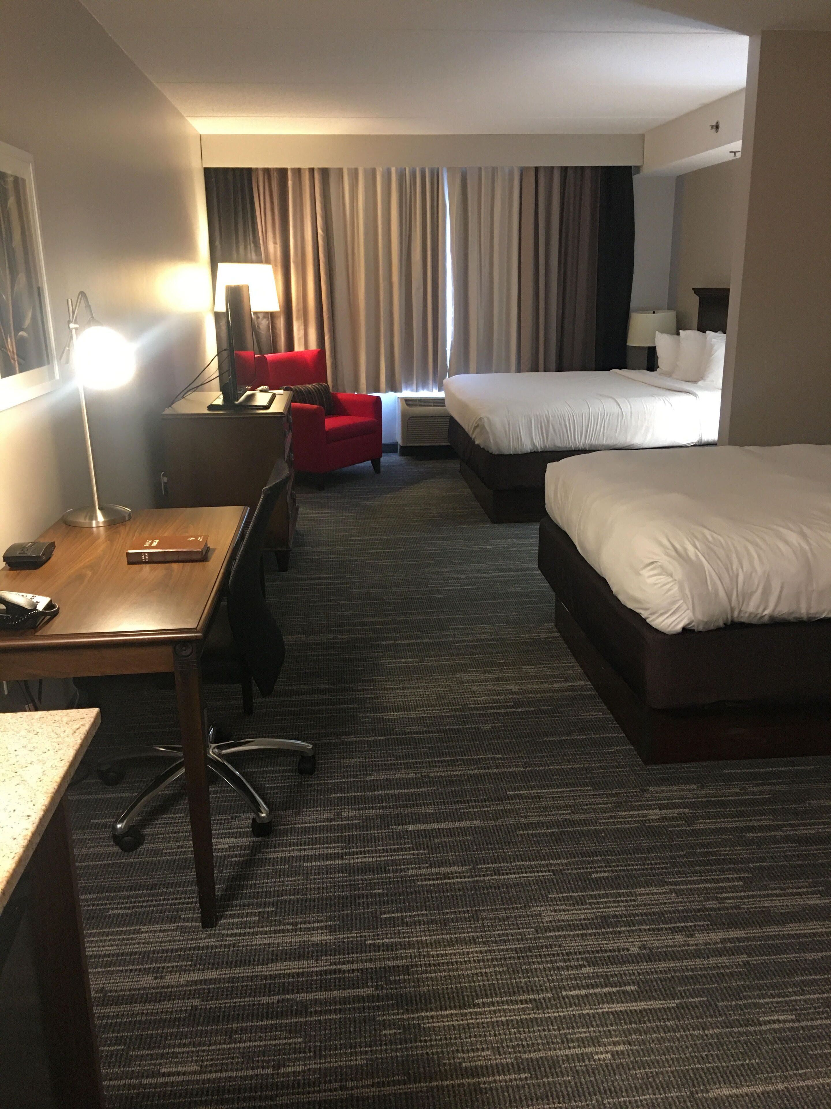 Ein Hotelzimmer mit Schreibtisch, zwei Betten, einem roten Sessel, einer Stehlampe und einem Vorhangfenster.