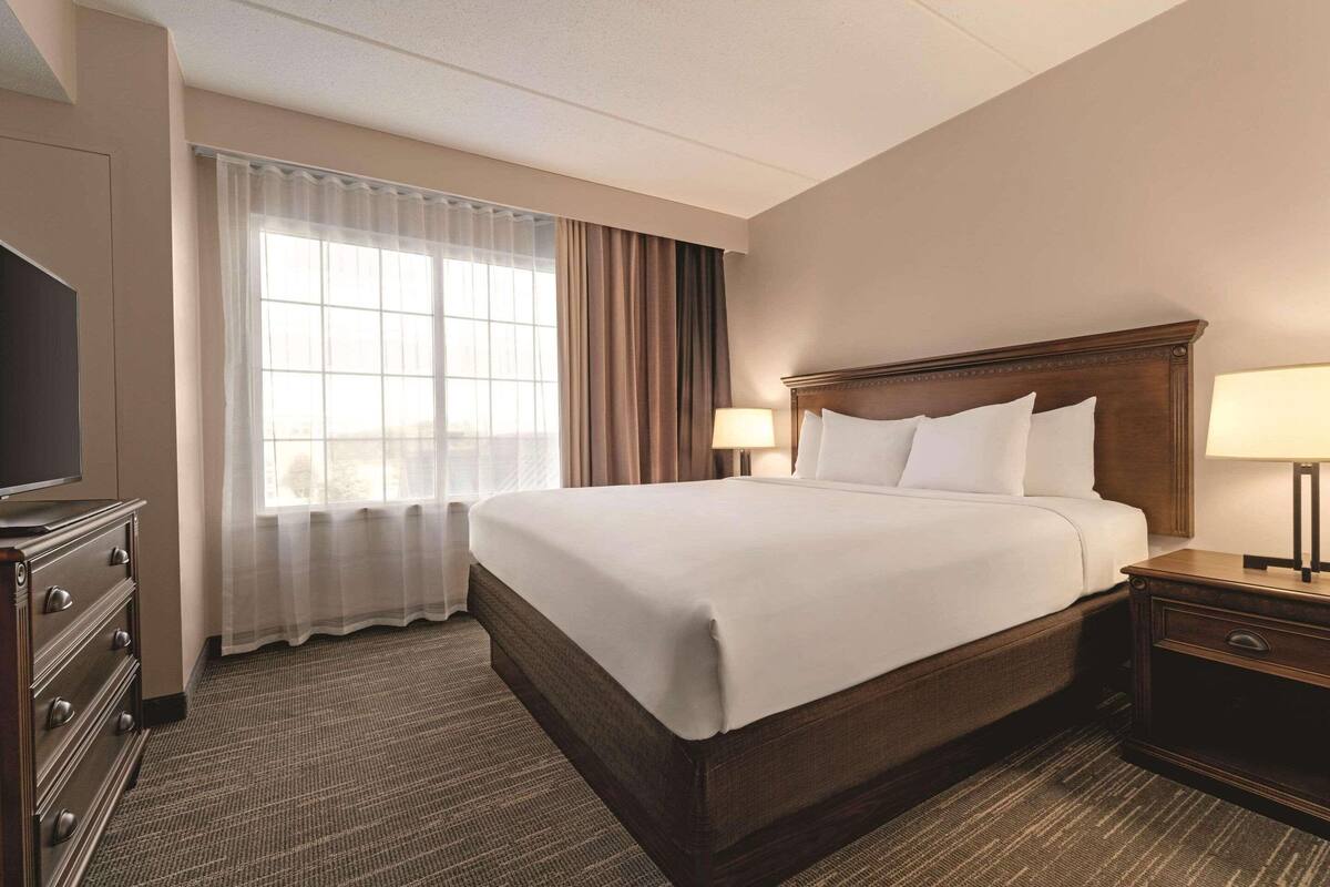 A hotel room with a large bed, two bedside tables with lamps, a TV on a dresser, and a window with curtains.