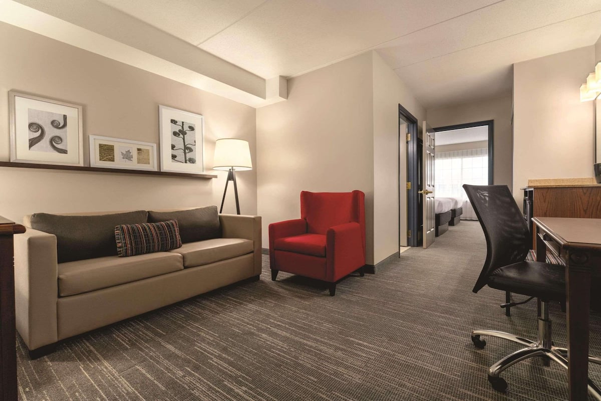 A hotel room with a sofa, a red armchair, a desk, and a lamp. There are framed pictures on the wall.