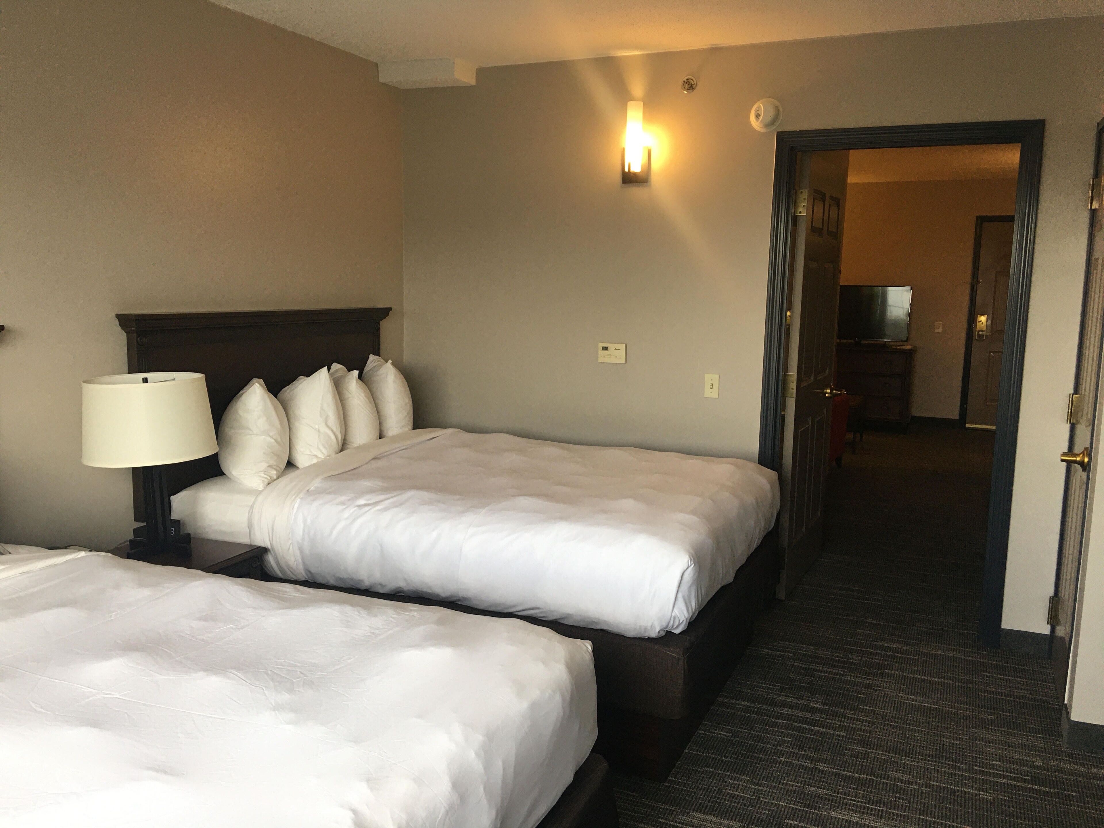 Une chambre d’hôtel avec deux lits, une table de chevet avec une lampe, un luminaire fixé au mur et une porte donnant sur une autre pièce.