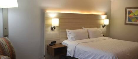 A hotel room with a bed, bedside lamps, a nightstand, and a patterned armchair.