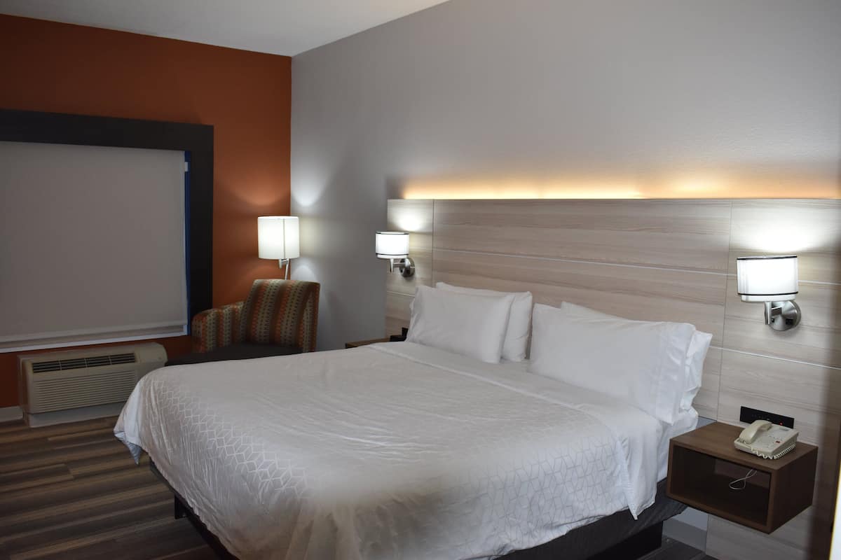 A hotel room with a large bed, two bedside tables with lamps, a projector screen, and a small seating area with a chair.