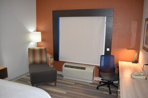 Una habitación de hotel con cama, escritorio con teléfono, aire acondicionado, una silla y una lámpara.