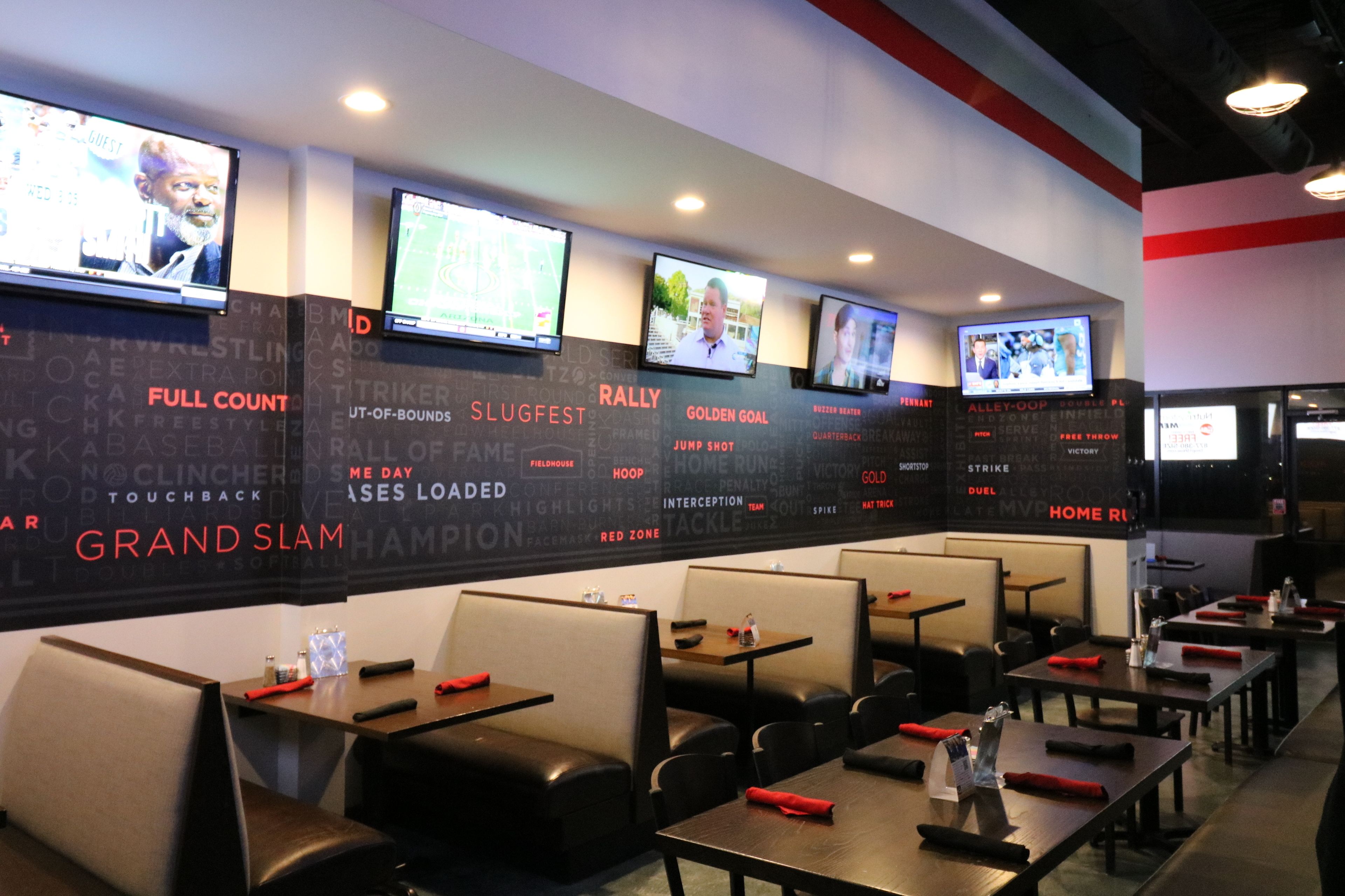 Un restaurante con temática deportiva que muestra contenido deportivo en varias pantallas, con cabinas y mesas adornadas con servilletas rojas.
