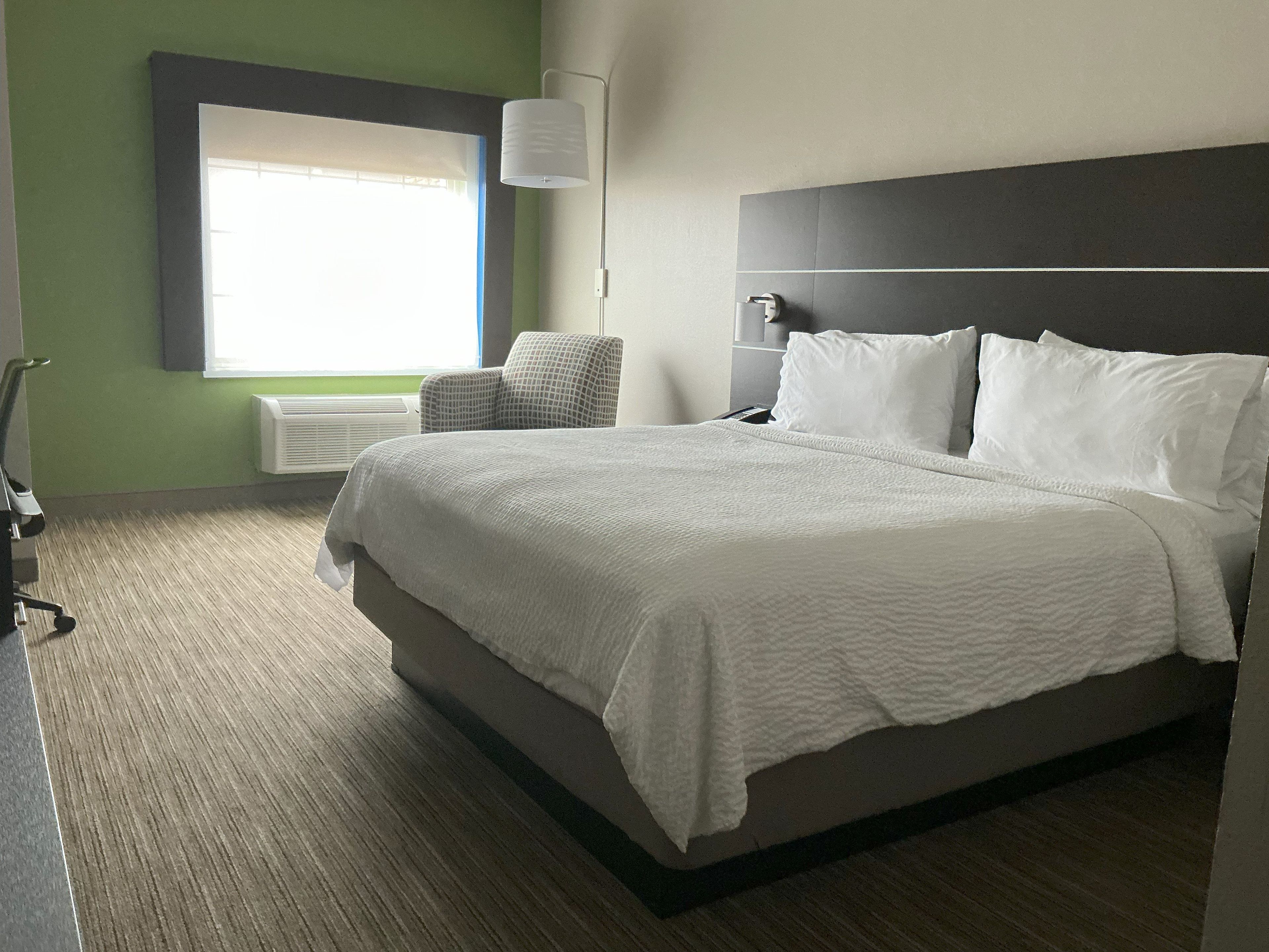 Quarto de hotel com uma cama, uma cadeira, uma janela e uma luminária fixada na parede.