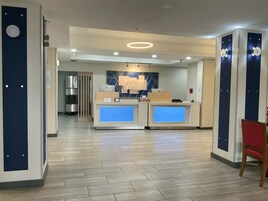 Zona de recepción de hotel con diseño moderno, que incluye un mostrador de recepción, una pantalla digital y una cocina al fondo.