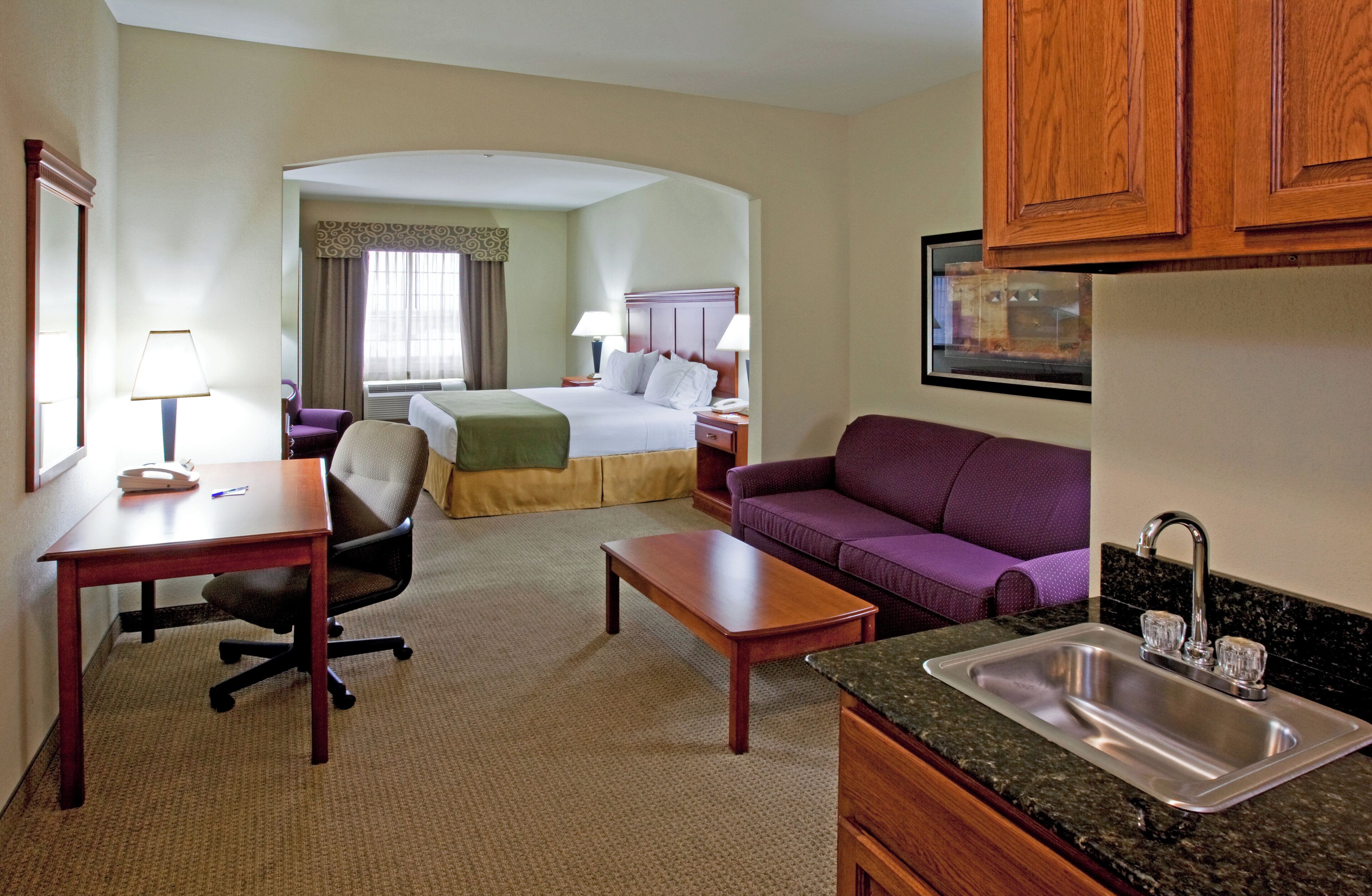 A hotel room with a bed, a sofa, a desk, a chair, a small table, a sink, and a window.