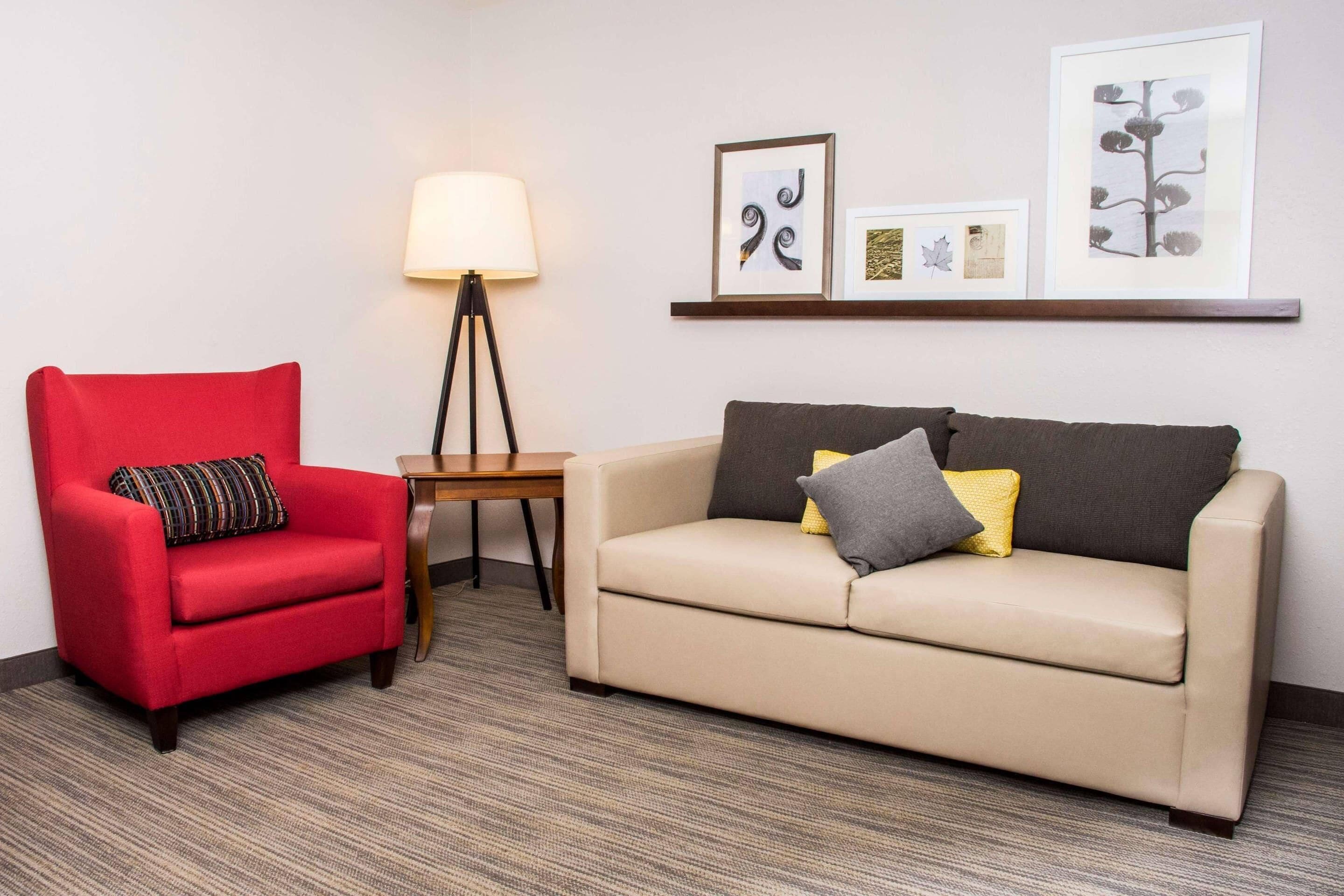 A room with a red armchair, a beige sofa, a small table, a floor lamp, and framed artwork on the wall.