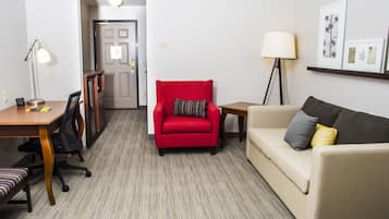 A hotel room with a red armchair, a beige sofa, a desk with a lamp, and a framed picture on the wall.