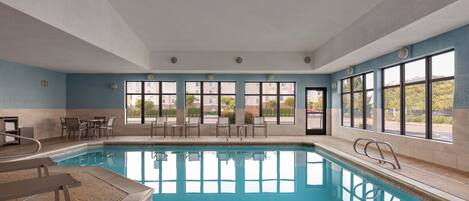 A large indoor swimming pool with clear blue water, surrounded by multiple windows offering views of the outside.