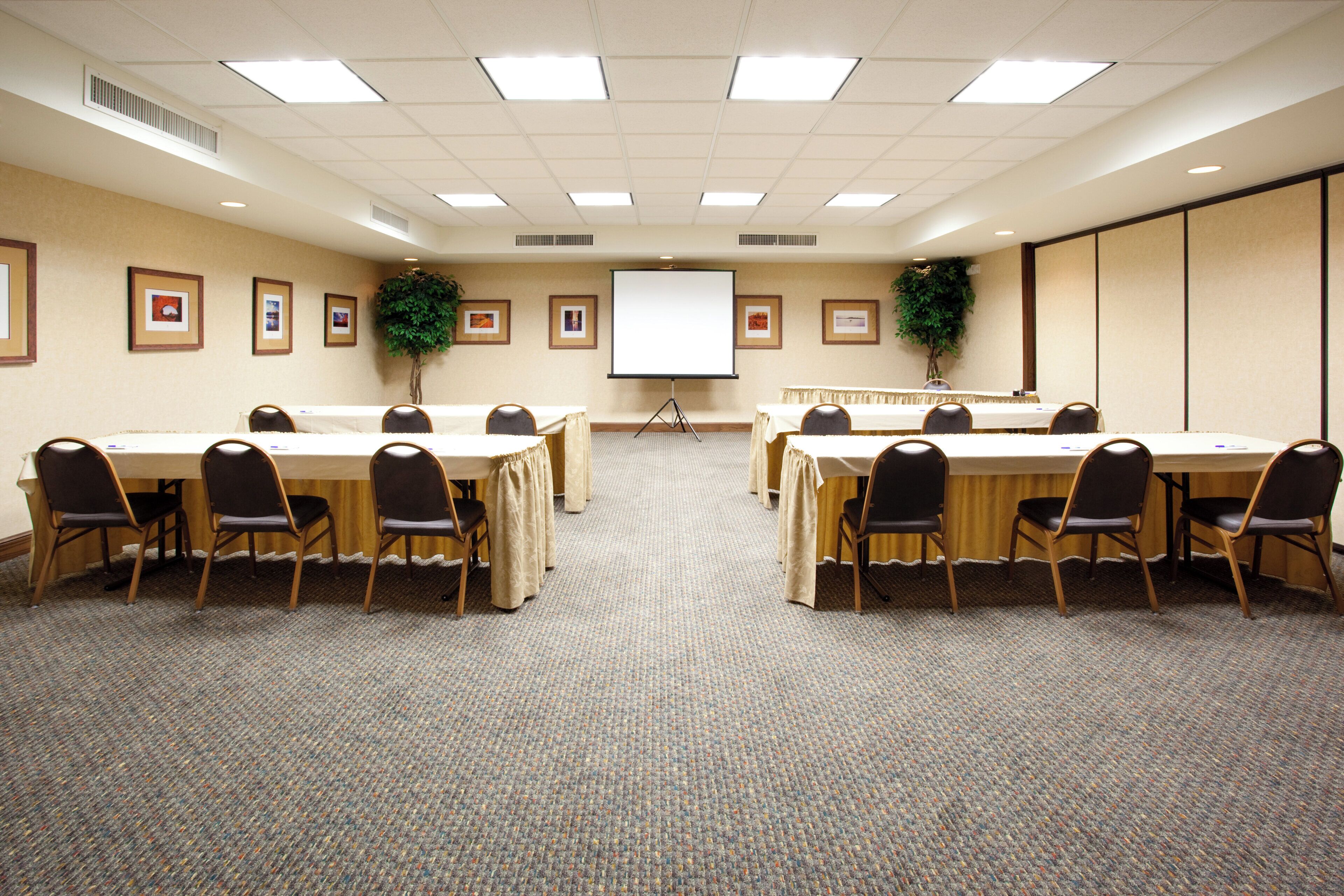 A conference room with two tables, chairs, a projector screen, and framed pictures on the walls.