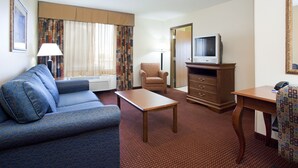A hotel room with a blue sofa, a wooden coffee table, a brown armchair, a TV on a wooden stand, and a desk with a telephone.