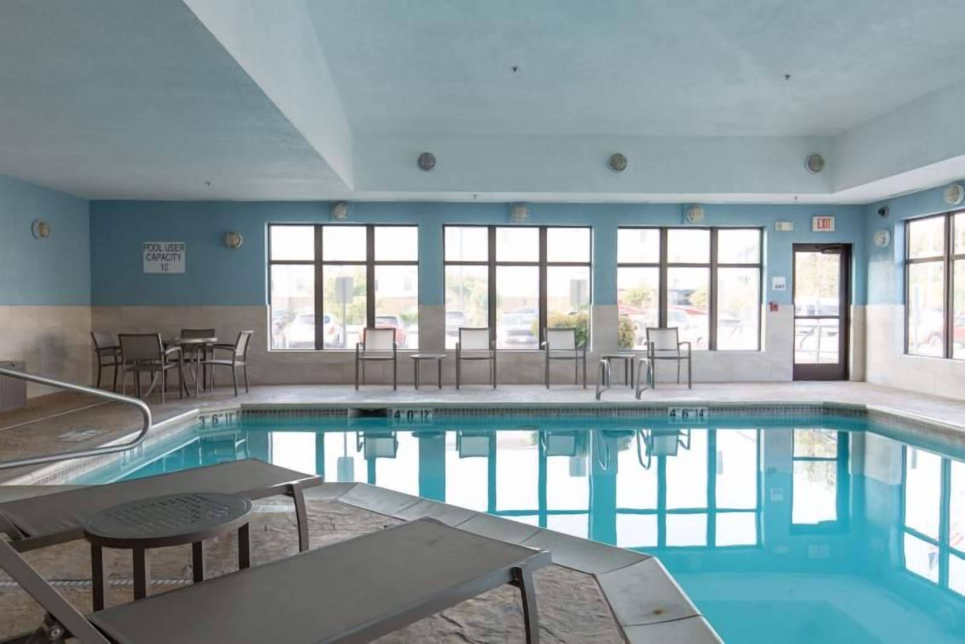 An indoor swimming pool with clear blue water, surrounded by windows, and equipped with sun loungers and tables.