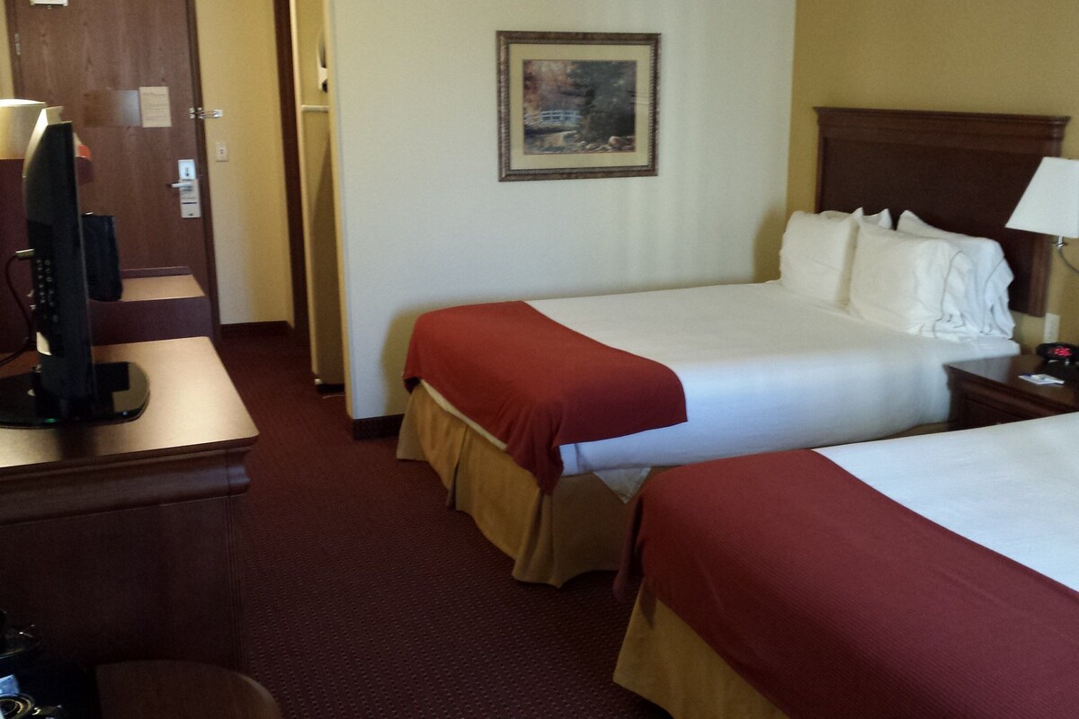 Quarto de hotel com duas camas, uma televisão, um quadro na parede e uma mesa de trabalho com abajur.