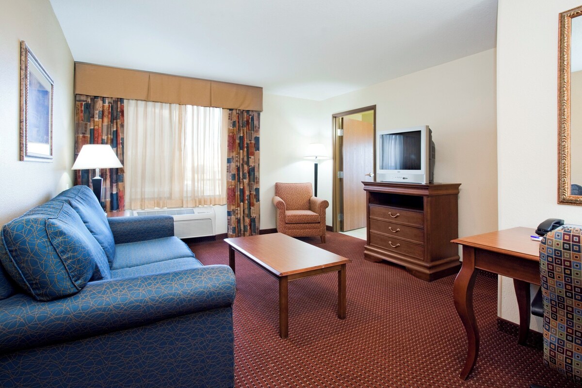 Quarto de hotel com um sofá azul, um suporte de TV de madeira, uma mesa e uma poltrona estampada.