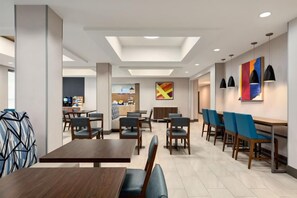 A modern hotel dining area with multiple tables and chairs, a visible kitchen area, and a colorful abstract painting on the wall. - Holiday Inn Express Hotel & Stes Salt Lake City-Airport East by IHG (Salt Lake City)