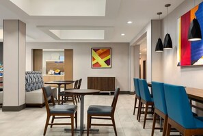 A modern dining area with tables and chairs, a kitchen in the background, and a colorful abstract painting on the wall.