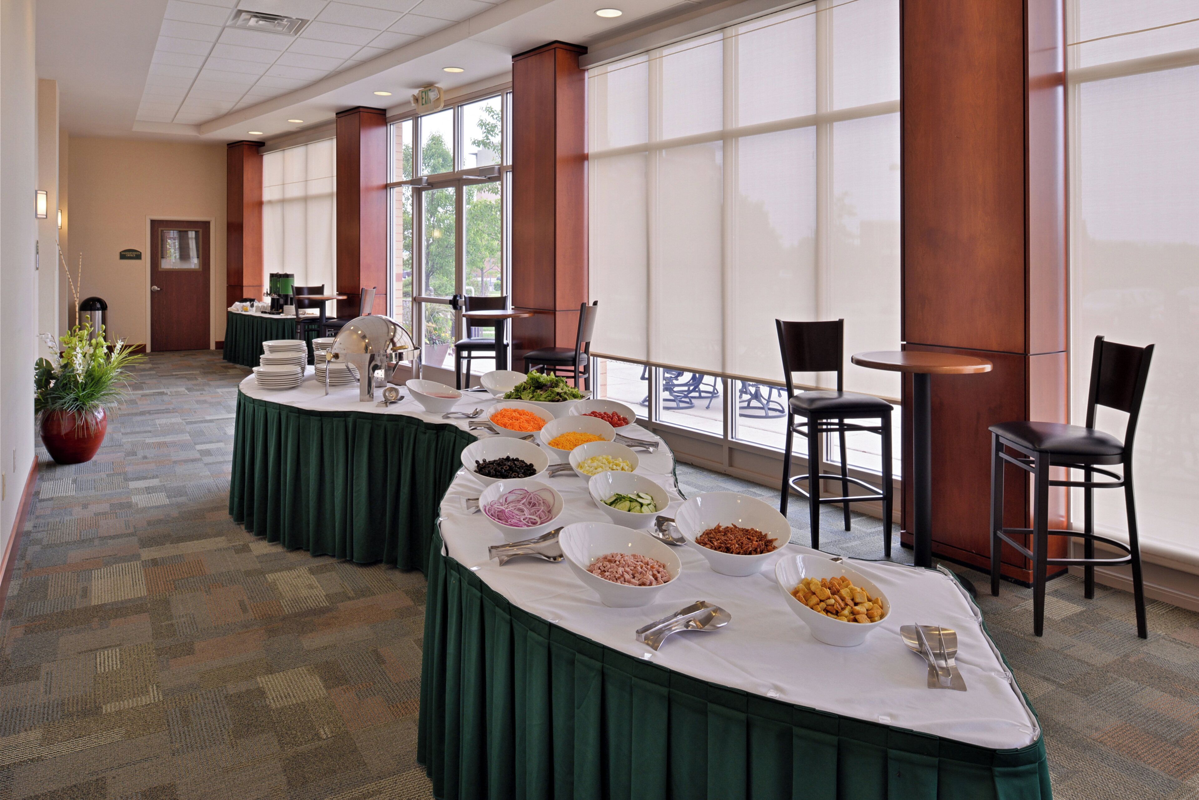 A buffet setup with various food items and utensils, a green tablecloth, and a view of the outdoors through large windows.
