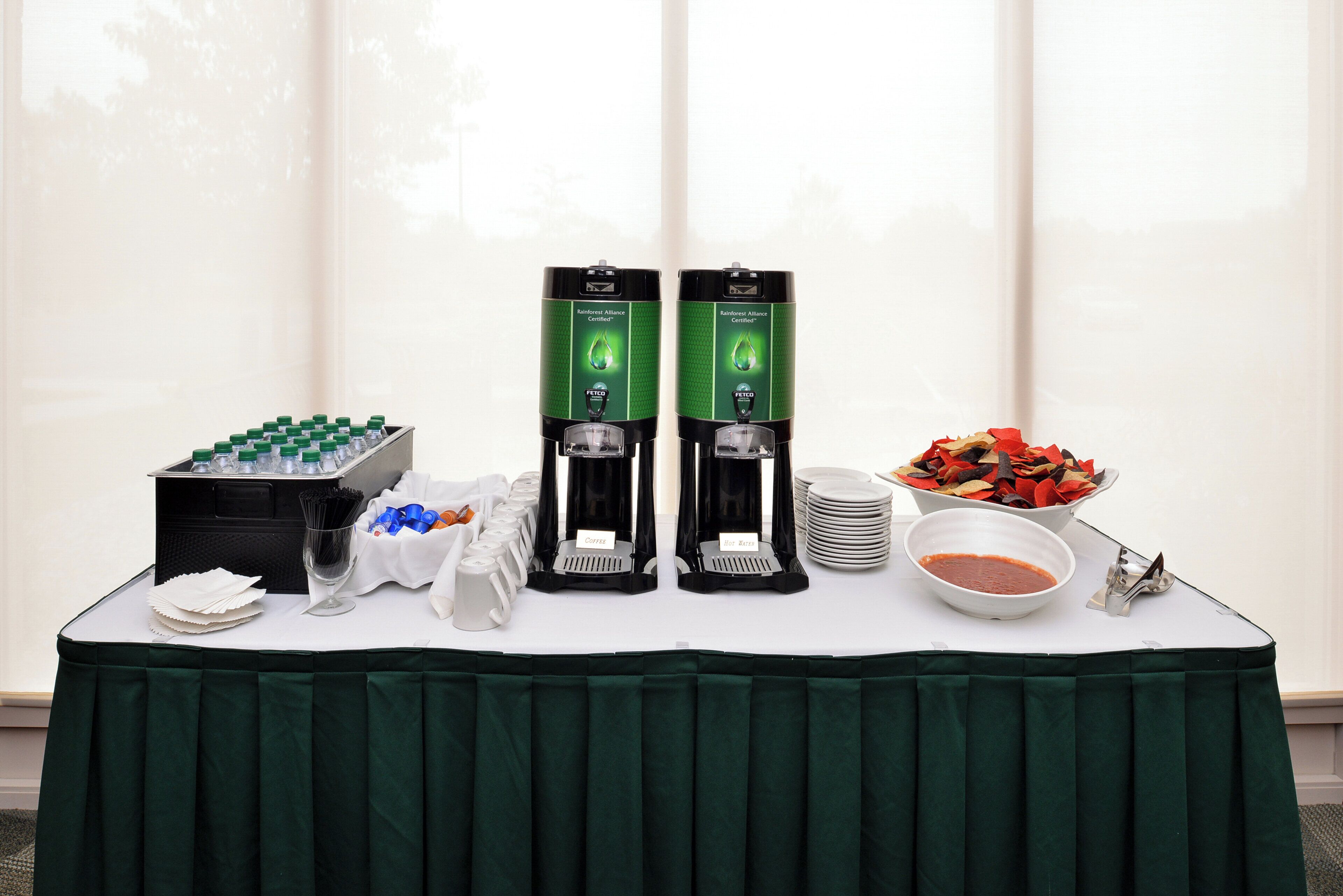A buffet table with two coffee dispensers, a water dispenser, a bowl of salsa, and a container of chips.